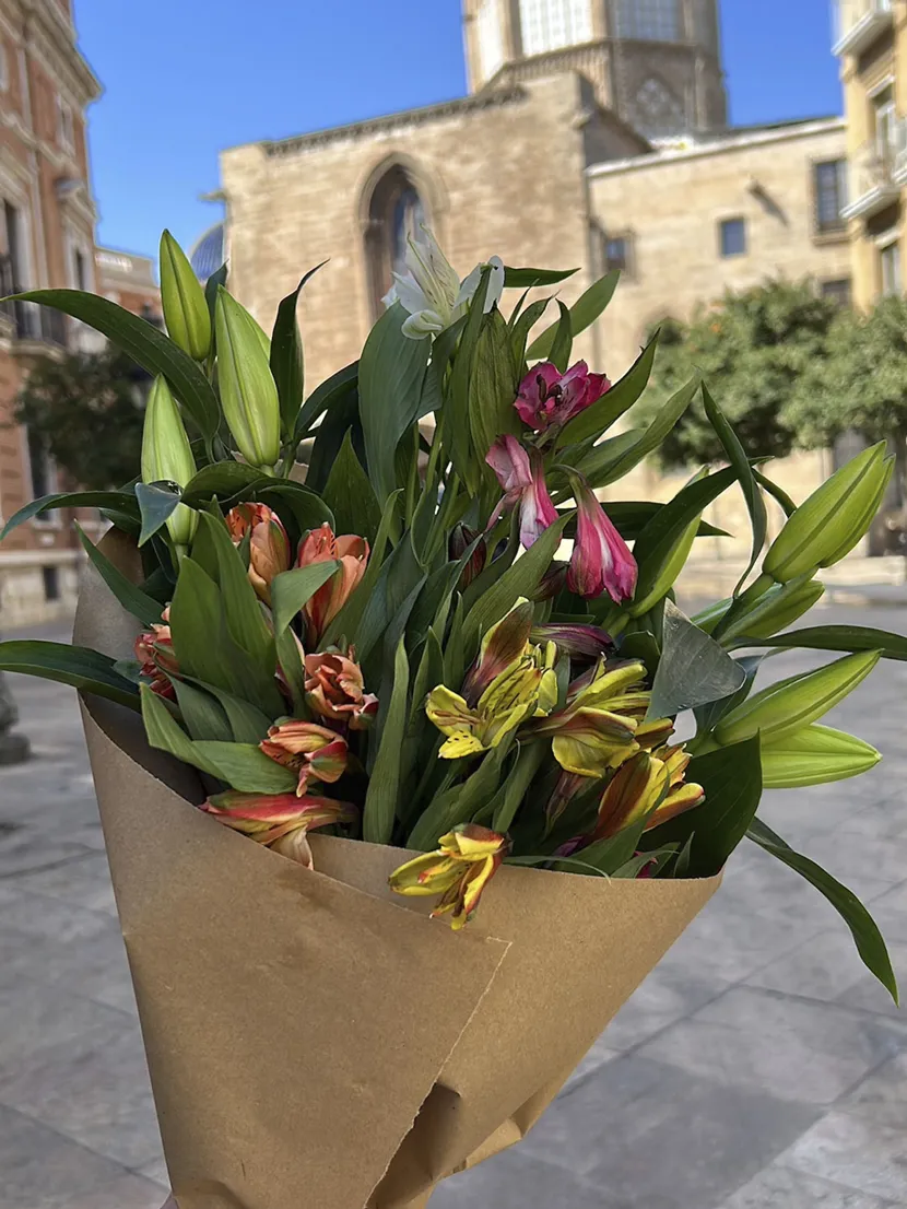 En hånd holder opp en fargerik blomsterbukett pakket inn i gråpapir. Blå himmel og spansk arkitektur i bakgrunnen.