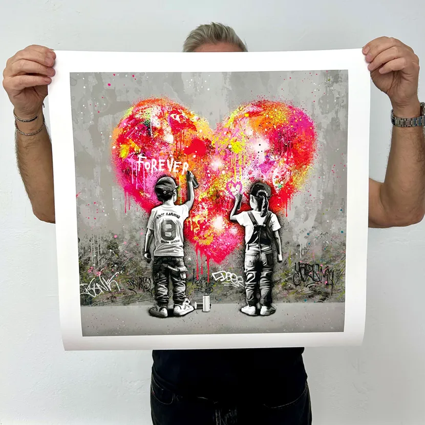 Kunsteren holder opp et trykk med motivet Forever U. Motivet består av et rosa, rødt og gult hjerte på en grå murvegg. En liten gutt og en jente i svart-hvitt maler på hjertet med spraybokser.