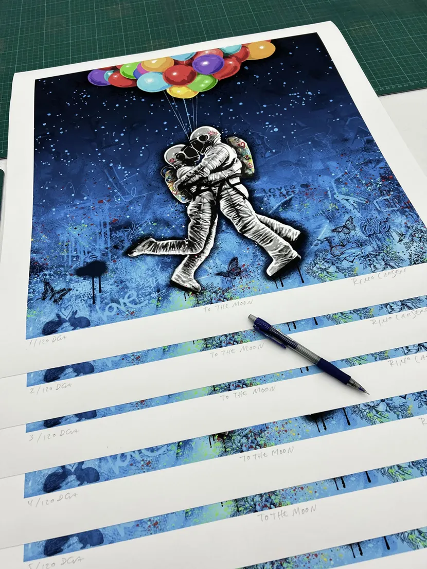 En stabel med trykk som signeres. Bildet heter To The Moon, motivet er to austronauter som svever under en samling fargerike ballonger.