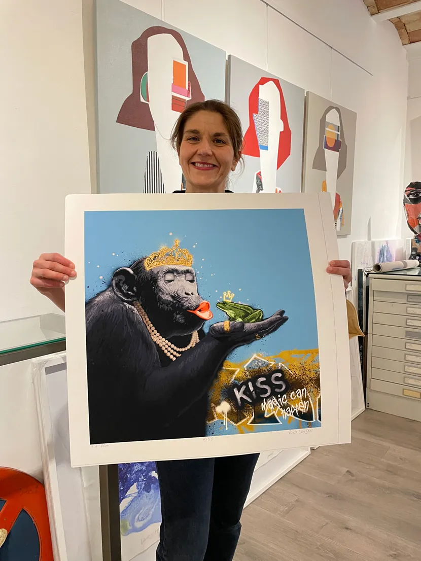 Kunstnerens spanske gallerist holder opp et trykk. Bildet heter Kiss B, motivet er en ape som kysser en frosk, på knall blå bakgrunn.